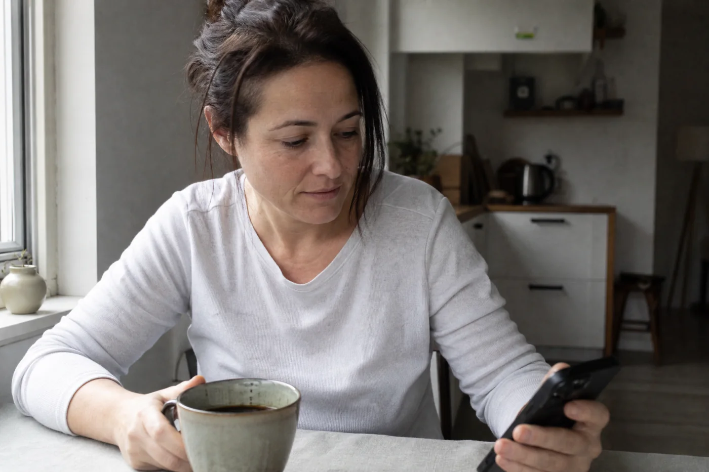 A woman at a kitchen table with a mug and her phone, morning light behind her.